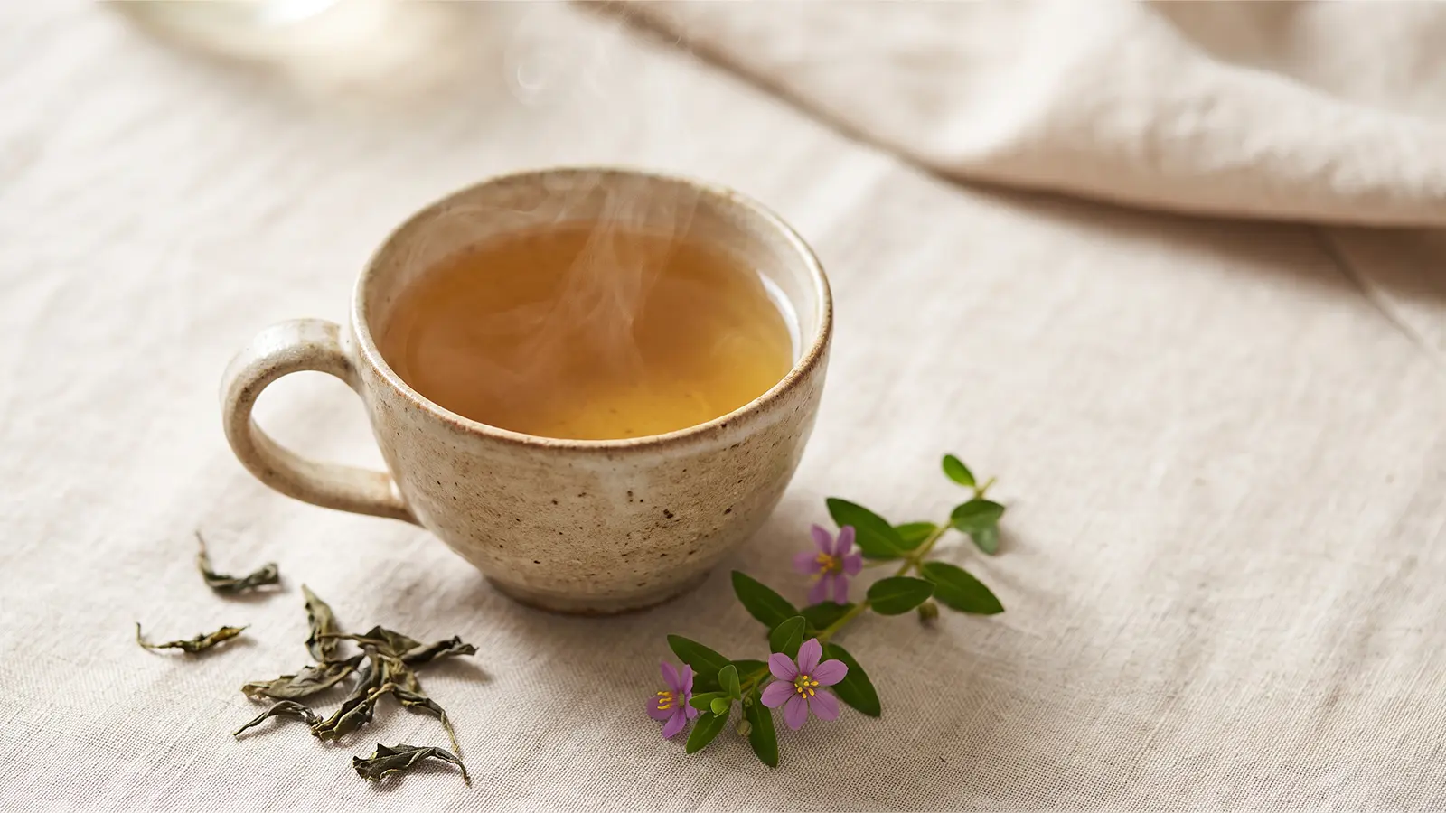 Cup of brewed Heen Bovitiya tea with rising steam, dried tea leaves, and a fresh purple Heen Bovitiya flower sprig